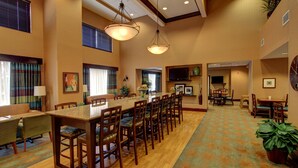 A spacious dining area with a long wooden table and multiple chairs, a television mounted on the wall, and a clock. - Hampton Inn Leesburg/Tavares (Leesburg)