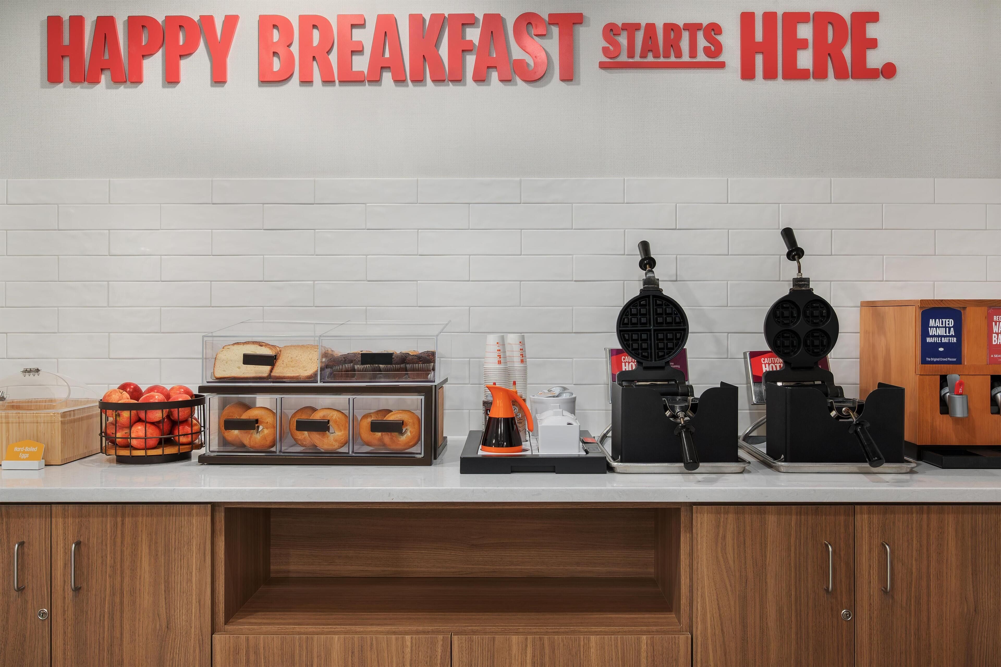 Una barra de desayuno con un letrero que dice “HAPPY BREAKFAST STARTS HERE”. Incluye una cafetera, una máquina para hacer gofres y una vitrina con muffins.