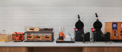 Una barra de desayuno con un letrero que dice “HAPPY BREAKFAST STARTS HERE”. Incluye una cafetera, una máquina para hacer gofres y una vitrina con muffins.