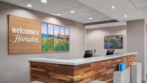 A reception desk with a wooden panel design, a "Welcome to Hampton" sign, and a "Leesburg, FL" poster. - Hampton Inn Leesburg/Tavares (Leesburg)