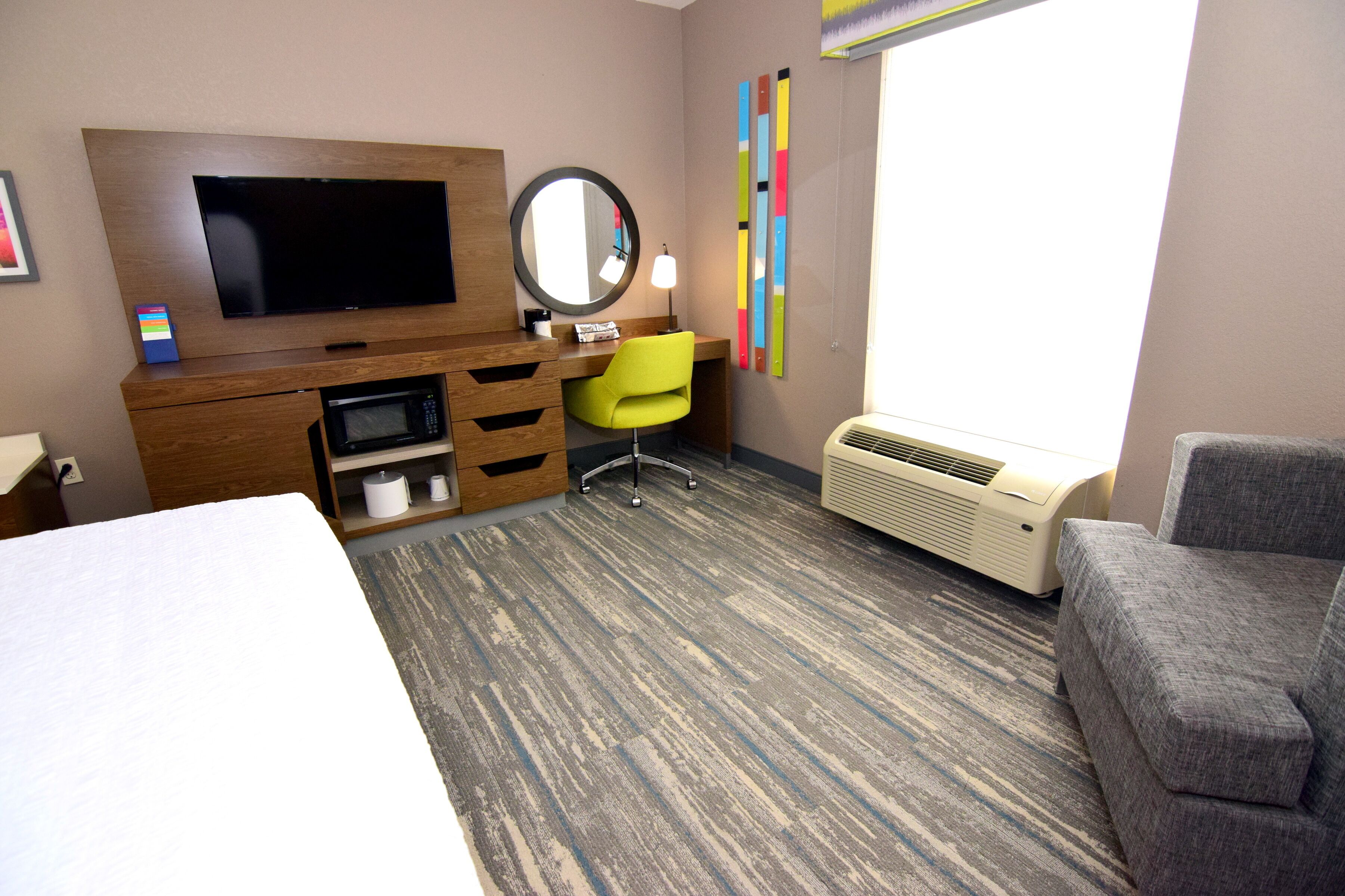 A hotel room with a bed, a desk, a chair, a television, a mirror, a heater, and a window.