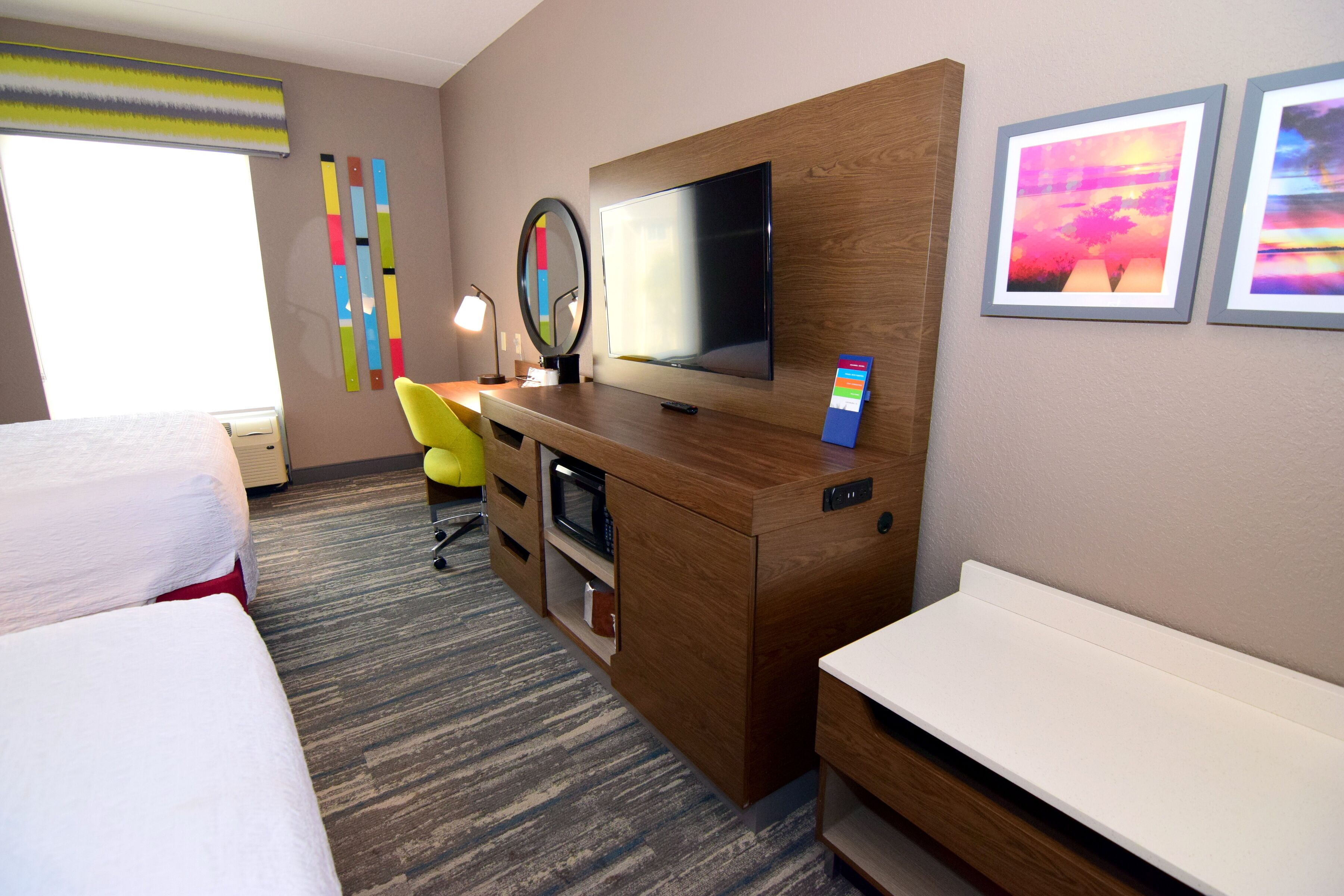 A hotel room with a bed, a desk with a computer, a TV, and artwork on the wall.