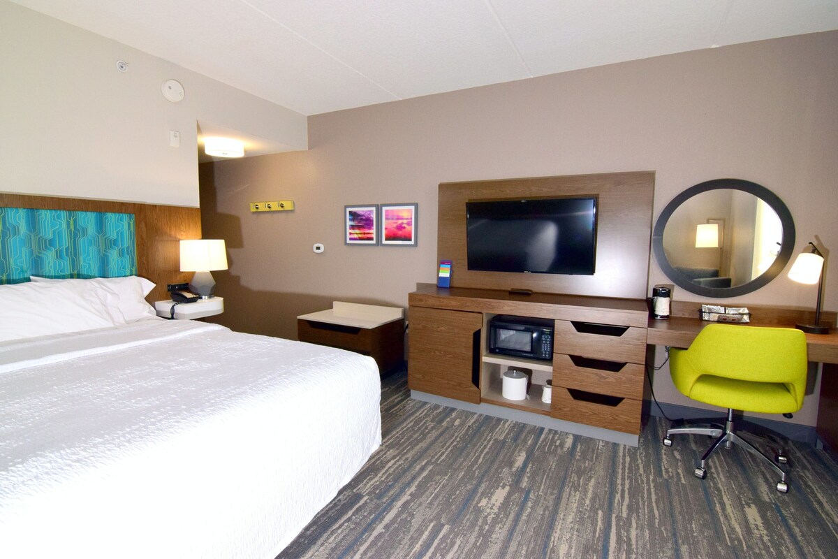 A hotel room with a bed, a desk with a chair, a television, a mirror, and a microwave.