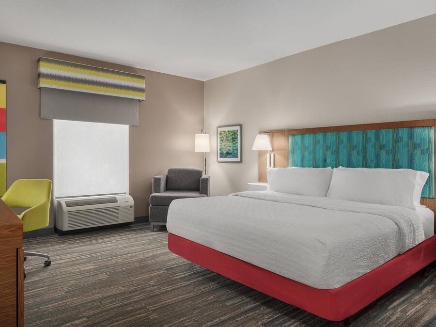A hotel room with a large bed, a desk, a chair, an air conditioning unit, and a window.