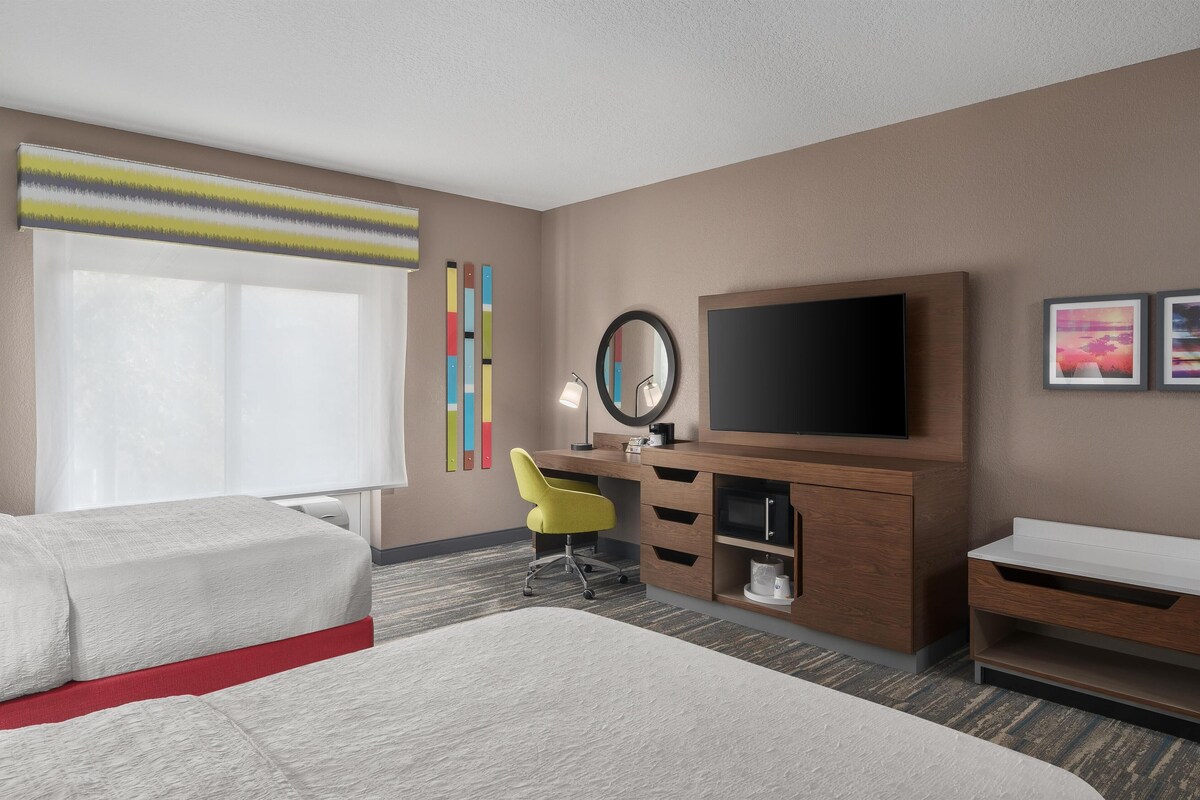 A hotel room with a bed, desk, chair, TV, and artwork on the wall.