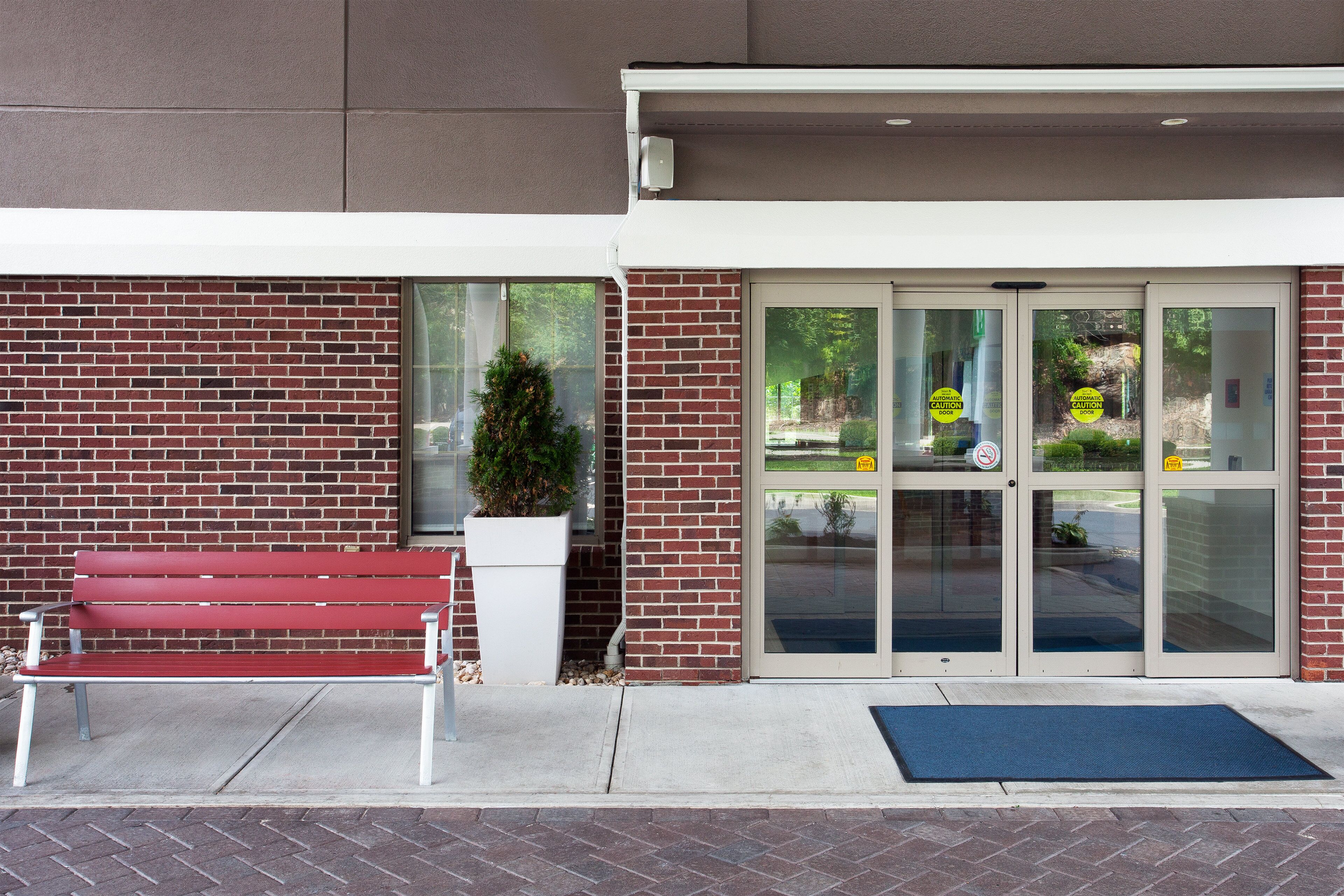 A building entrance with a red bench, a potted plant, and a blue mat.