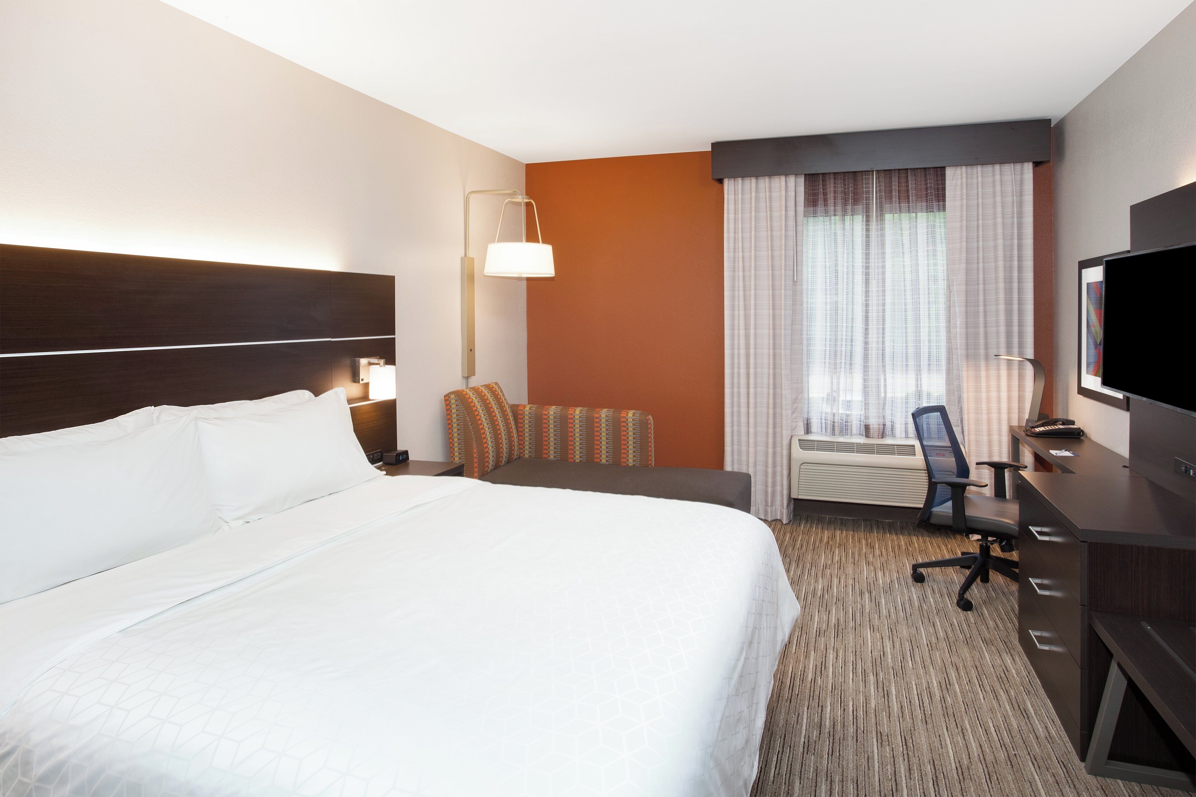 A hotel room with a large bed, a desk with a computer, a TV, and a window with curtains.