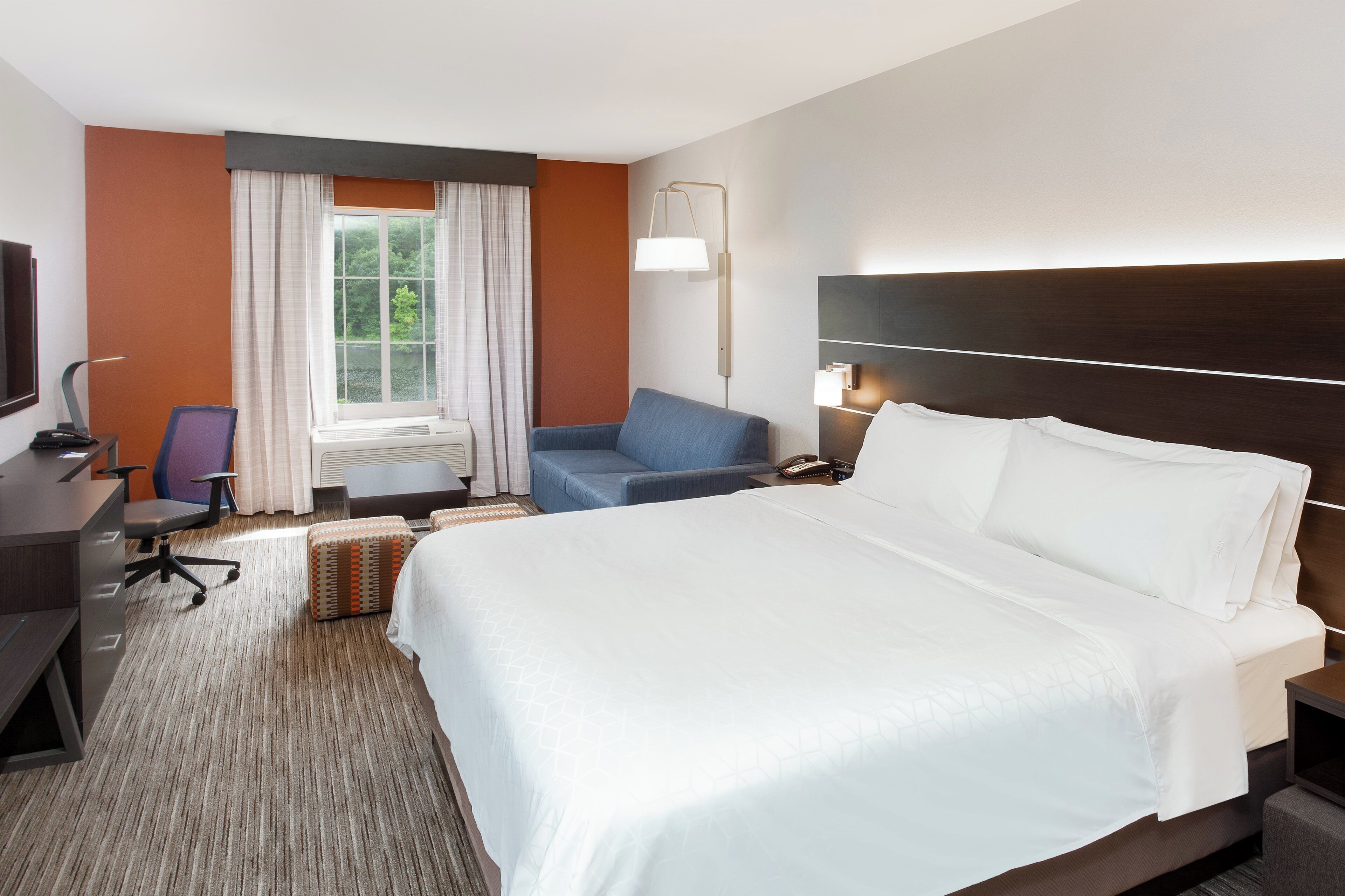 Habitación de hotel con una cama grande, un escritorio con computadora, una silla y un ventanal con cortinas.