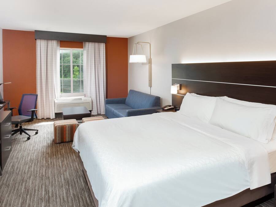 Habitación de hotel con una cama grande, un escritorio con computadora, una silla y un ventanal con cortinas.