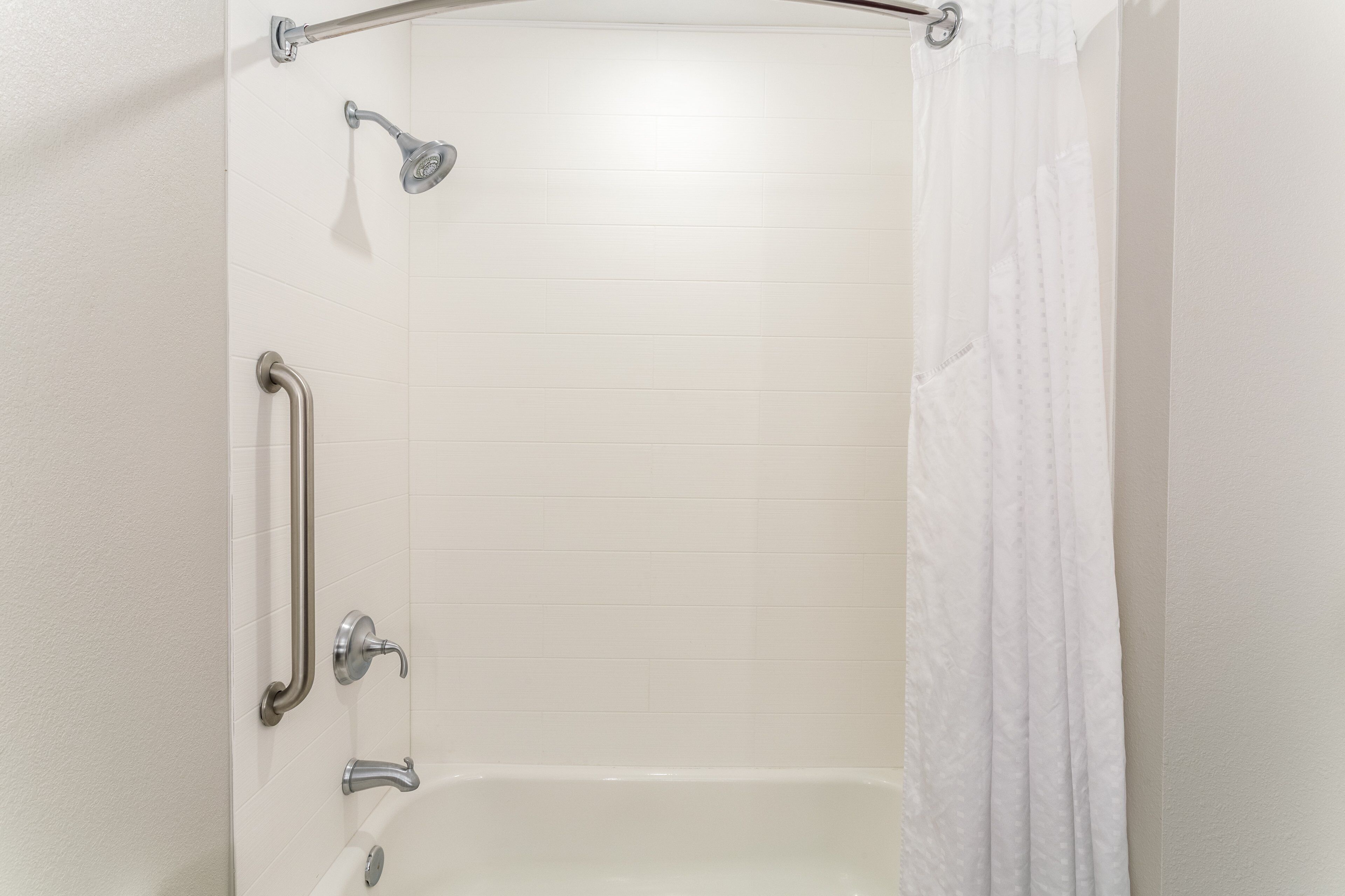 Ein Badezimmer mit weiß gefliesten Wänden, einer Dusche mit Duschbrause, einer Haltestange und einer Badewanne.