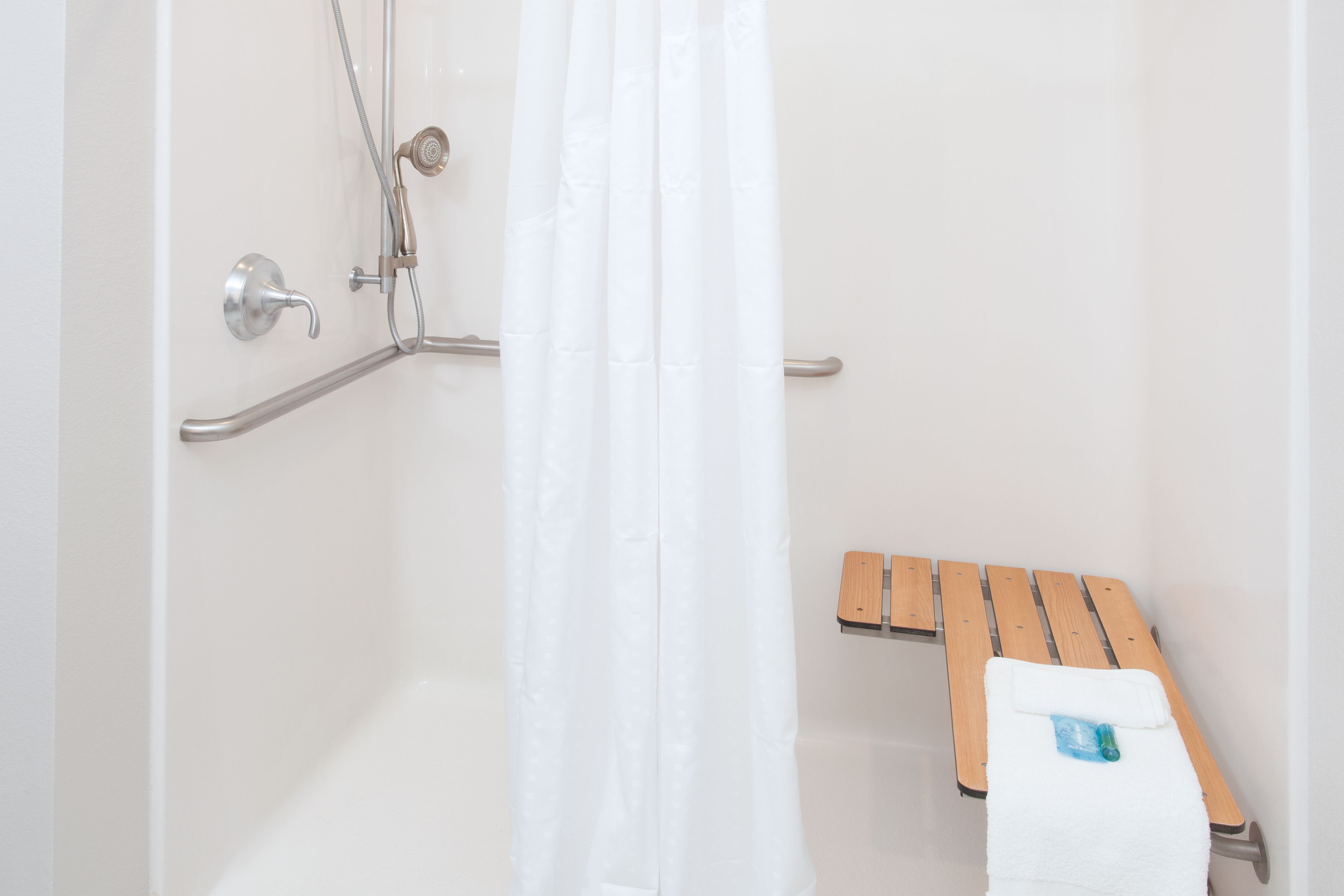 A shower area with a white shower curtain, a handheld showerhead, and a wooden bench with a towel.