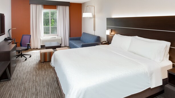 A hotel room with a large bed, a desk with a chair, a sofa, a lamp, and a window with curtains. - Holiday Inn Express Haskell - Wayne Area by IHG (Wanaque)