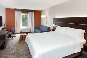 Quarto de hotel com uma cama grande, uma escrivaninha com cadeira, um sofá, um abajur e uma janela com cortinas.