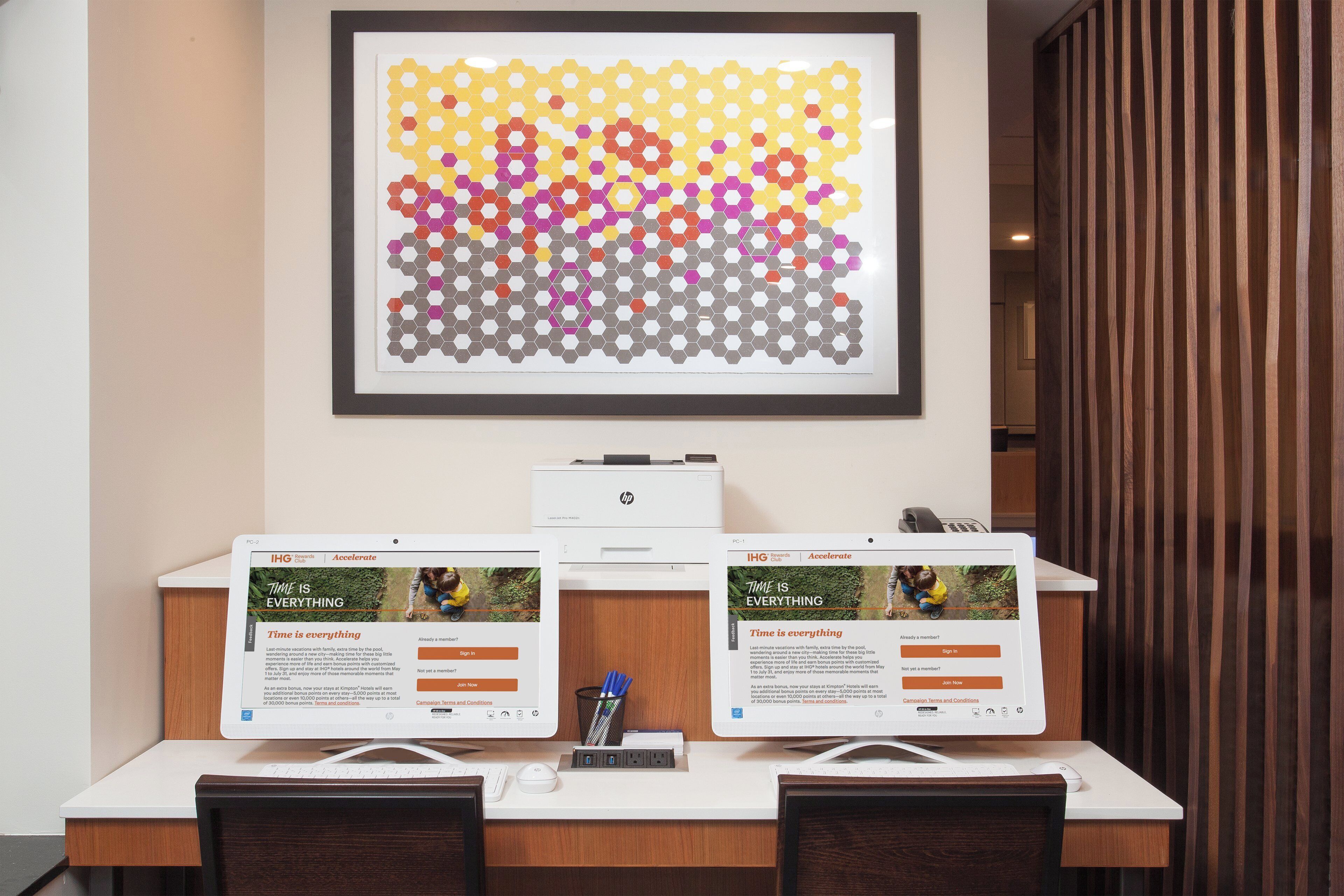 A desk with two HP LaserJet printers, a framed abstract art piece, and two computer monitors displaying a hotel rewards program webpage.