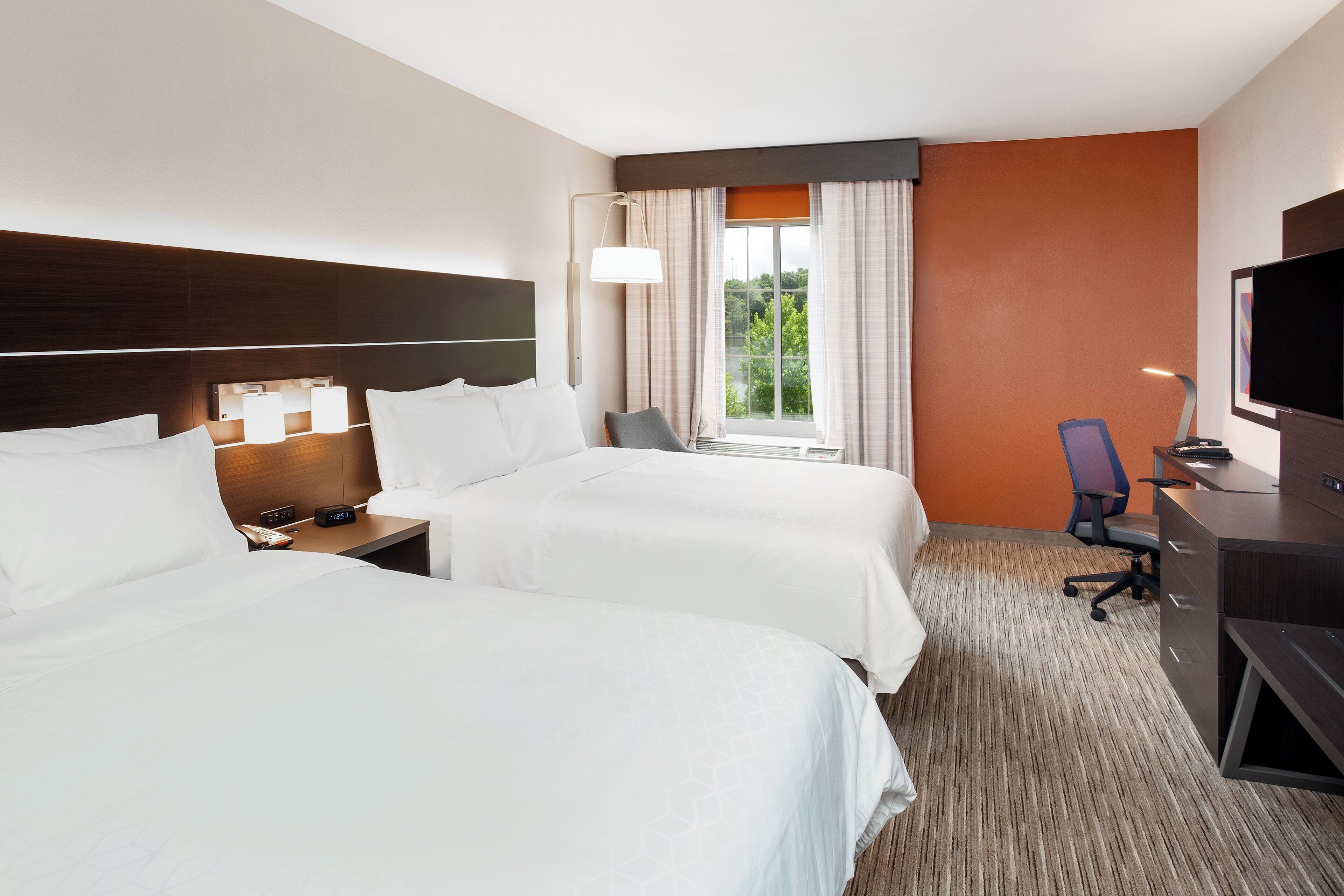 Habitación de hotel con dos camas, un escritorio, una silla, un televisor y un ventanal con cortinas.