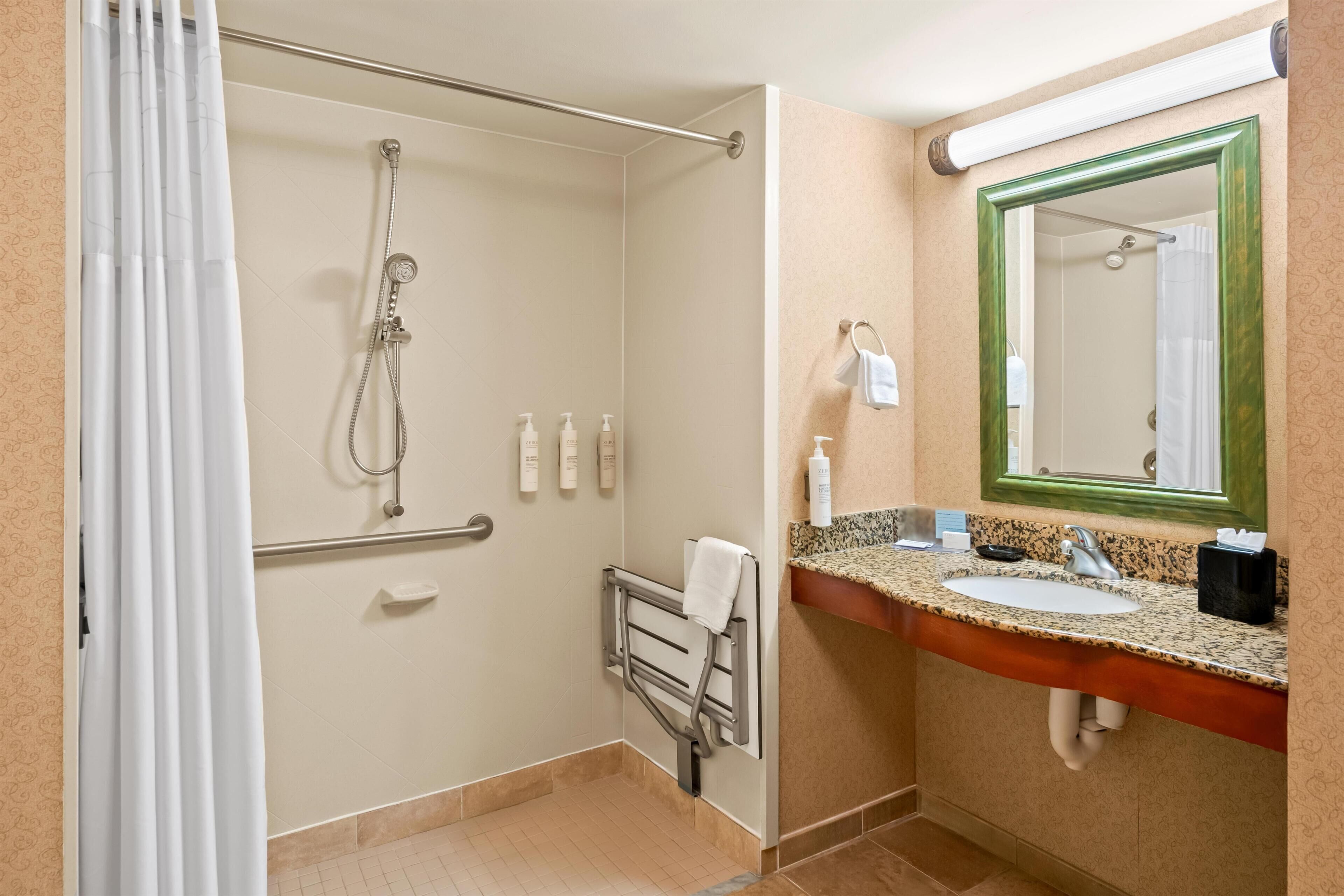 A bathroom with a shower, a handrail, a towel rack, a sink with a mirror, and toiletries.