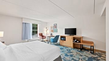 A hotel room with a bed, a desk with a TV, a chair, a small table, and a window with curtains.