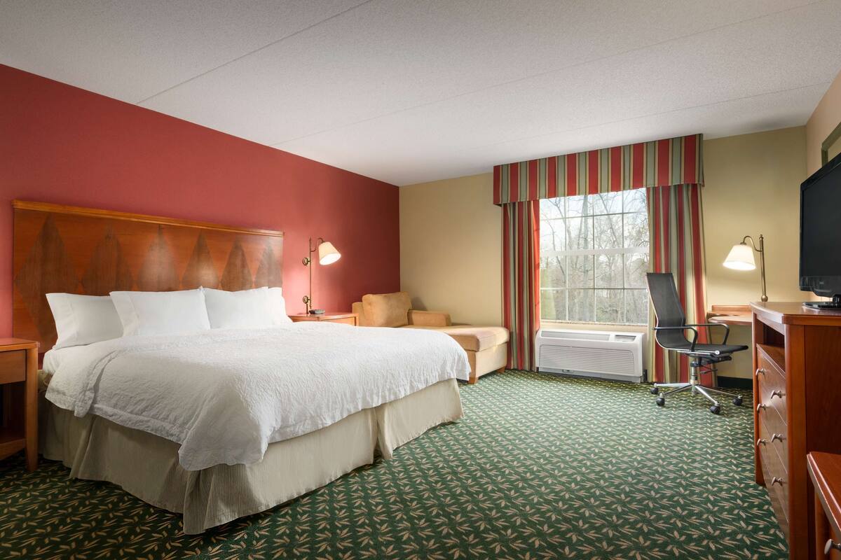 A hotel room with a large bed, a desk with a chair, a window with curtains, a radiator, and a television.