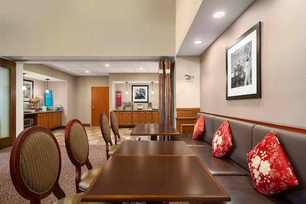 A hotel dining area with a long table, cushioned seating, and a kitchen in the background. - Hampton Inn & Suites Williamsburg-Central (Williamsburg)