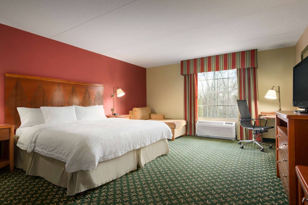 A hotel room with a large bed, a desk with a chair, a window with curtains, and a radiator.