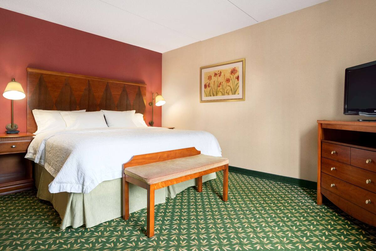 A hotel room with a bed, bedside lamps, a bench, a dresser, and a television.