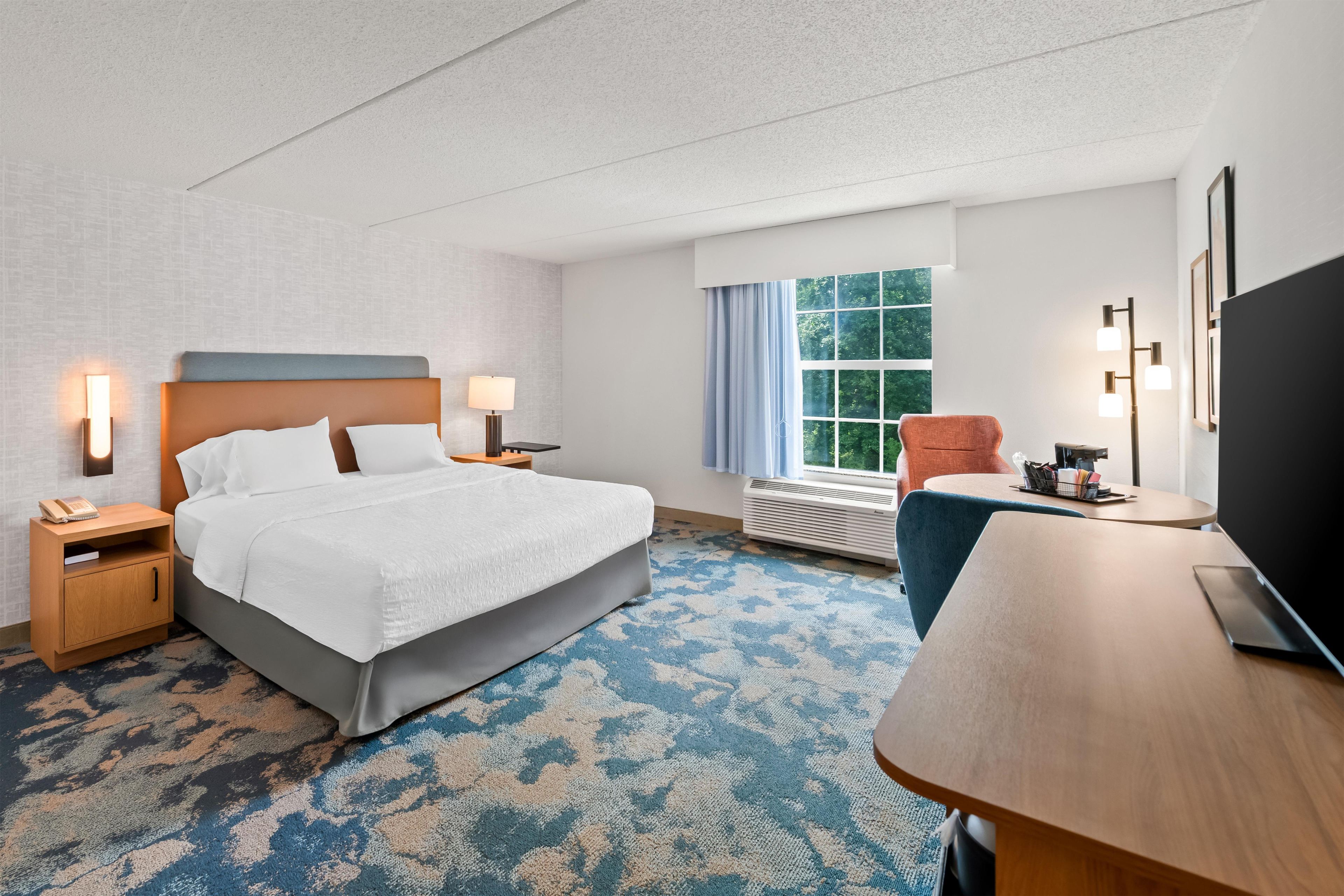 Quarto de hotel com cama, mesas de cabeceira, escrivaninha, cadeira, televisão e janela com cortinas.