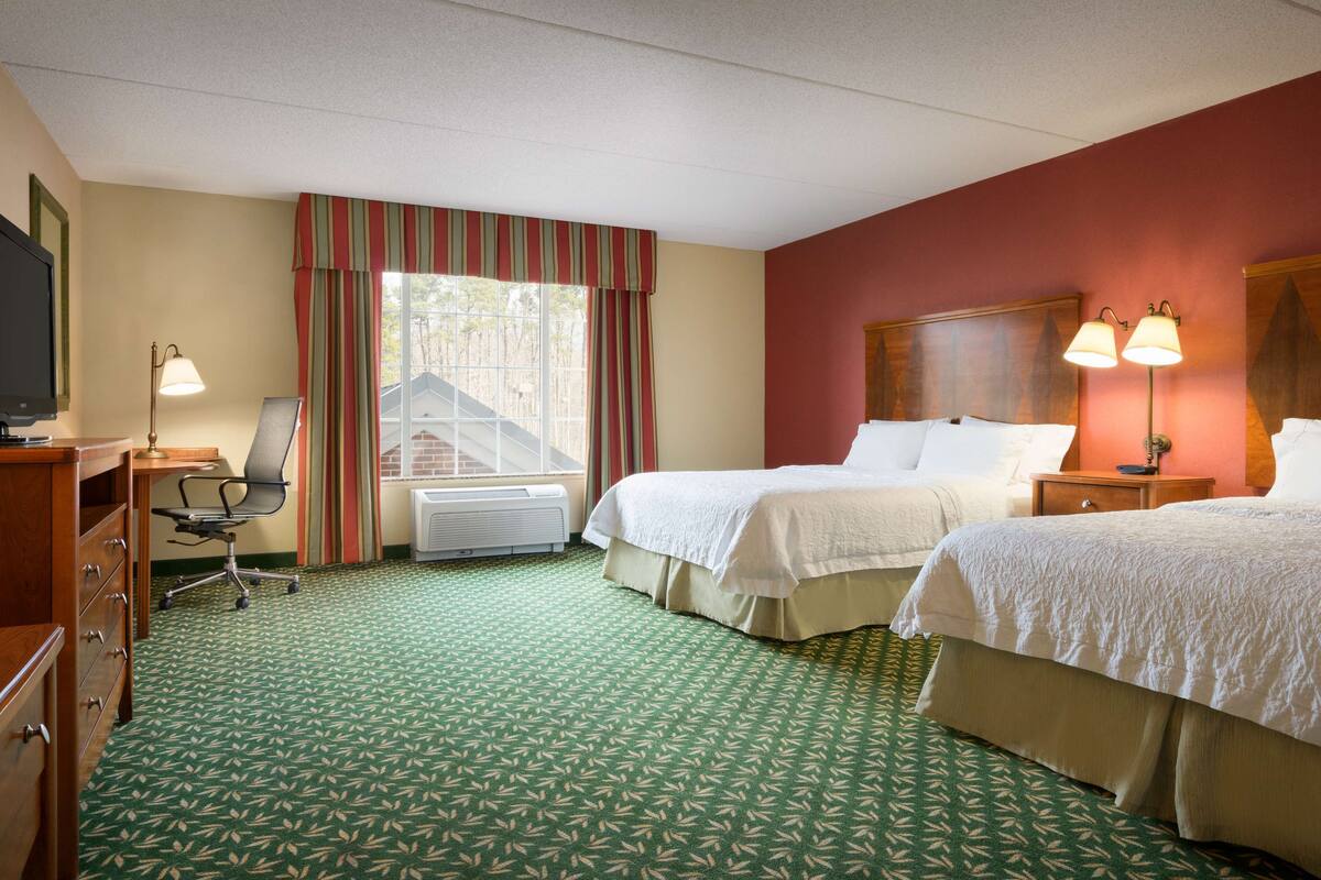A hotel room with two beds, a desk with a chair, a television, and a window with curtains.