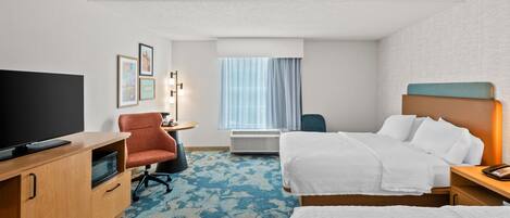 A hotel room with a flat-screen TV, a desk with a lamp, two beds, a chair, and a large window with curtains.