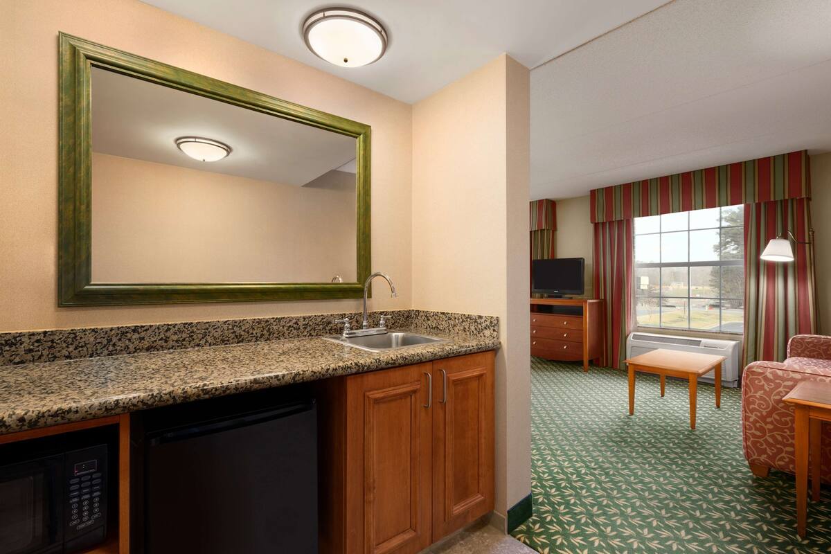 A compact hotel room with a kitchenette, a large mirror, a television, and a seating area with a patterned carpet.