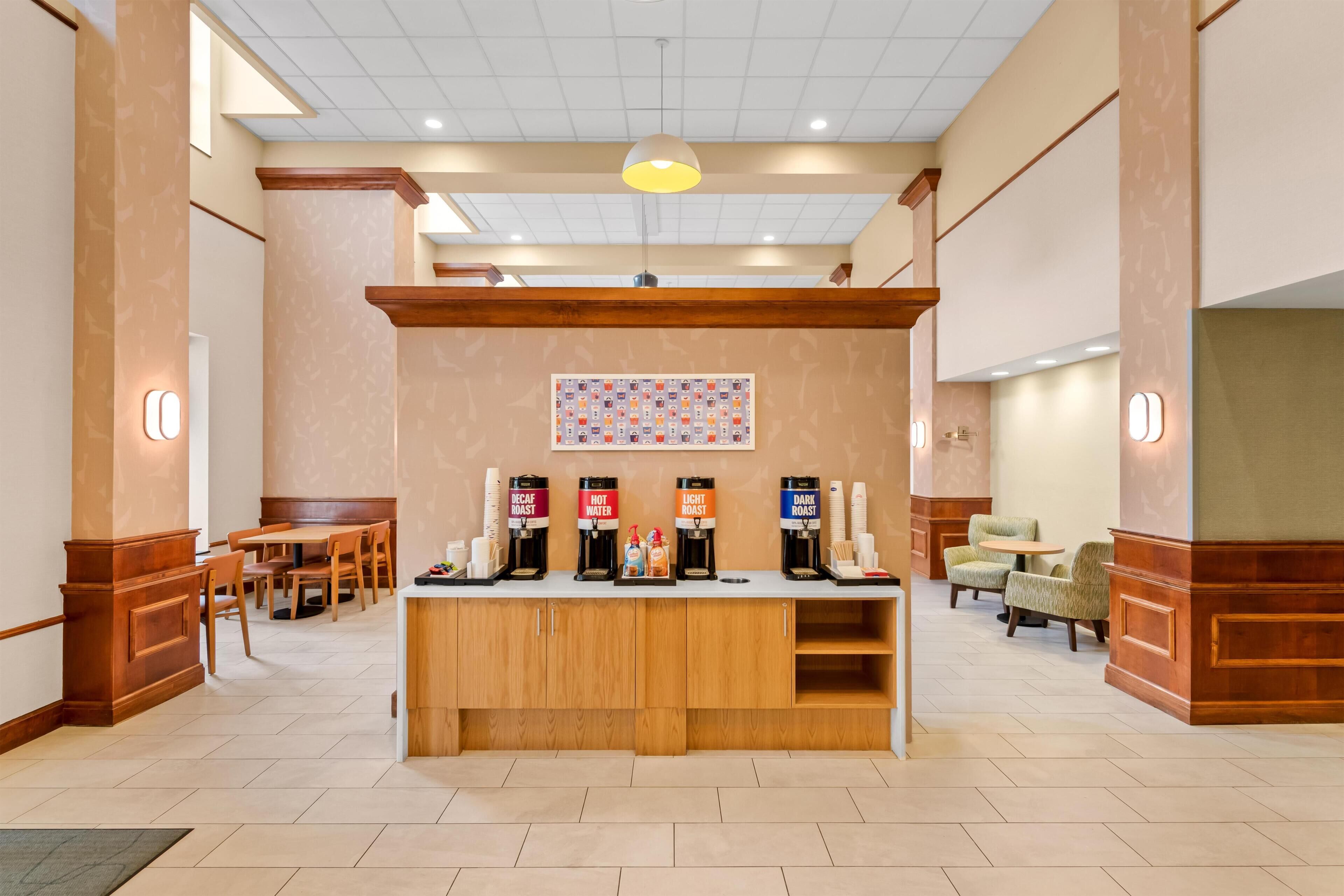 A hotel lobby with a coffee station, seating area, and a wall-mounted artwork.