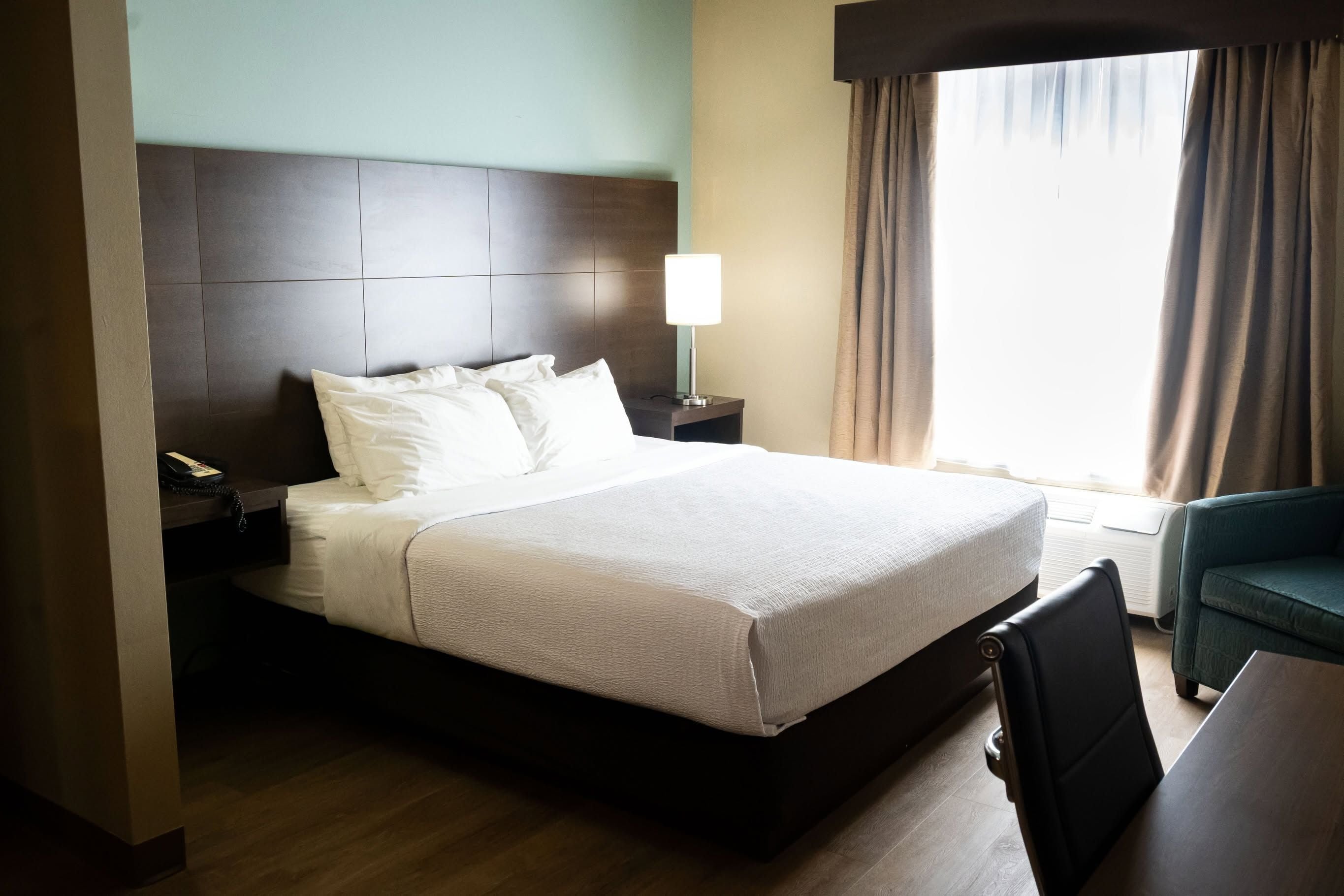 Una habitación de hotel con una cama grande, una mesita de noche con lámpara, una silla y un ventanal con cortinas.