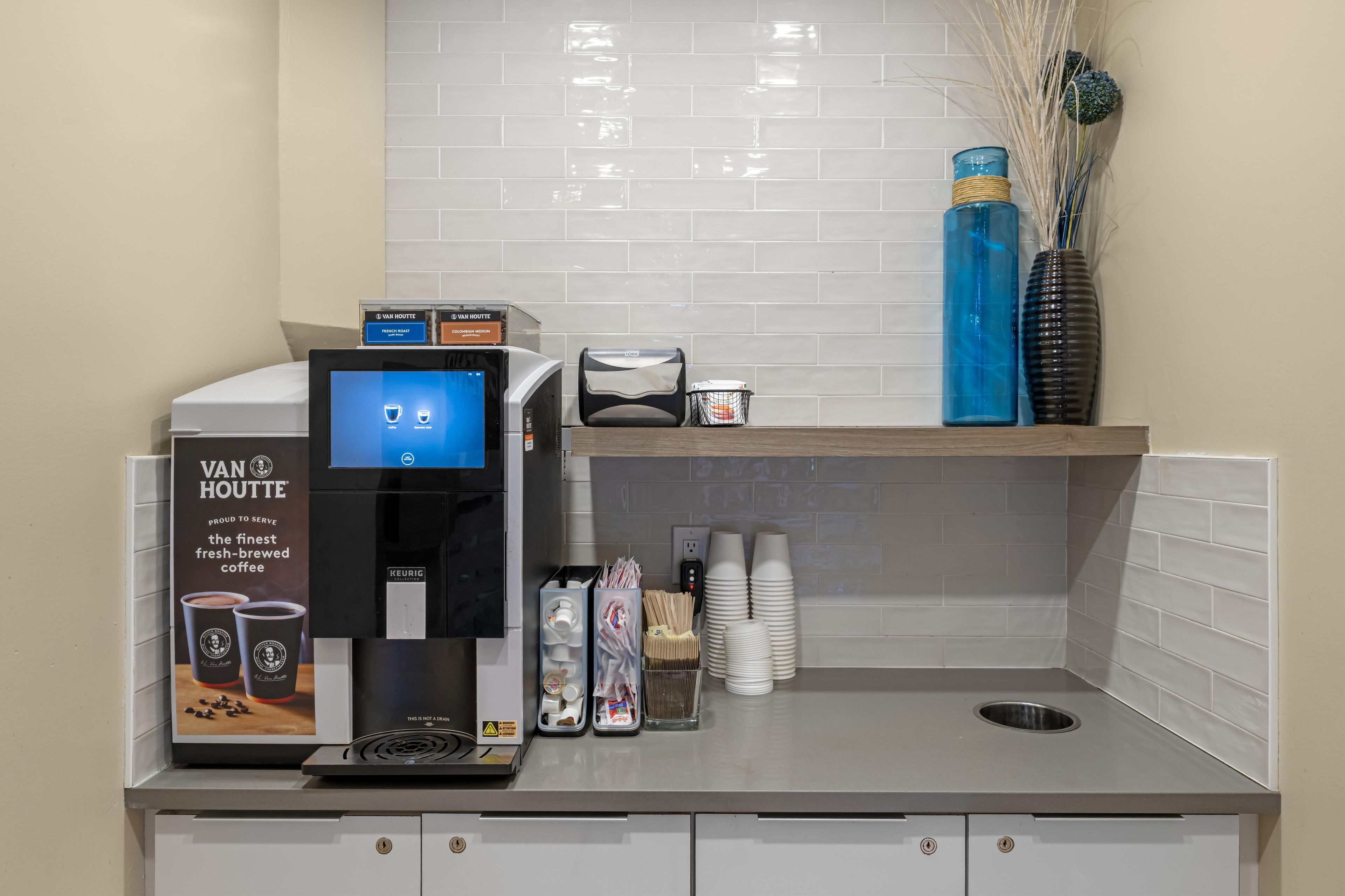 A coffee station with a Keurig machine, a Van Houtte coffee advertisement, and a small shelf with a vase and cups.
