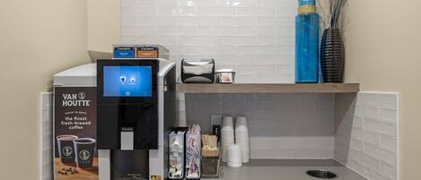 A coffee station with a Keurig machine, a Van Houtte coffee advertisement, and a small shelf with a vase and cups.