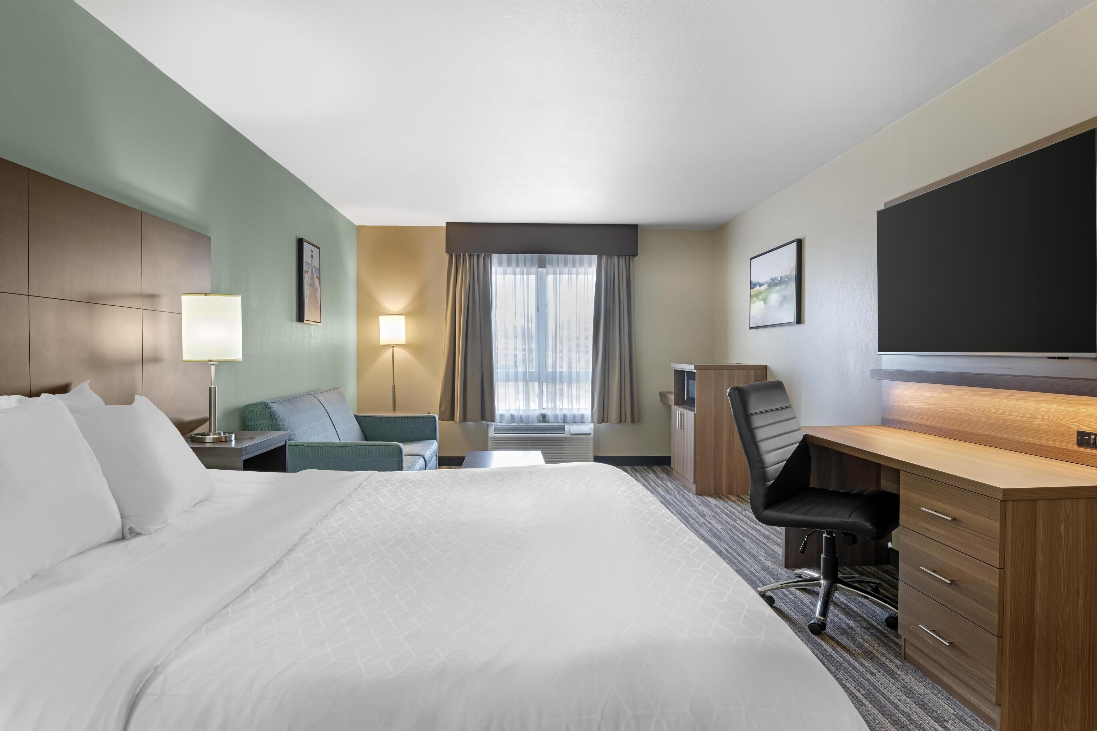 A hotel room with a large bed, a desk, a chair, a lamp, a TV, and a window with curtains.