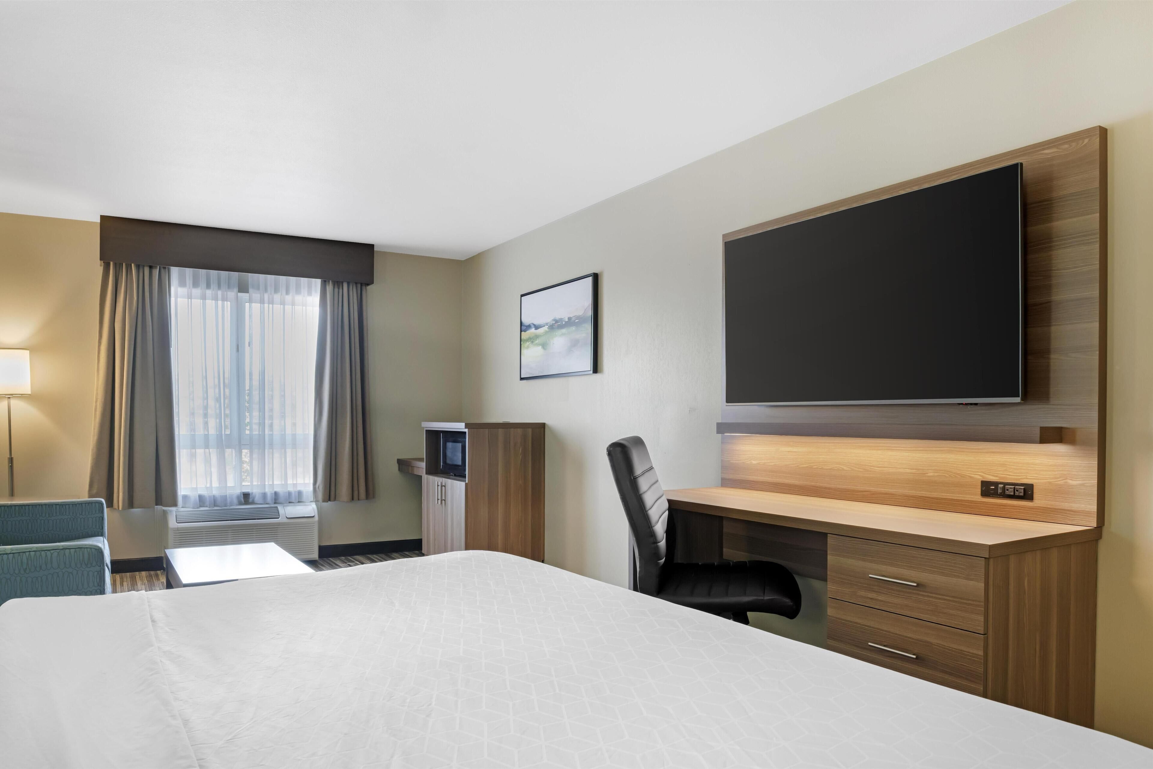 A hotel room with a bed, a desk, a chair, a TV, a desk lamp, a window with curtains, and a wall-mounted picture.