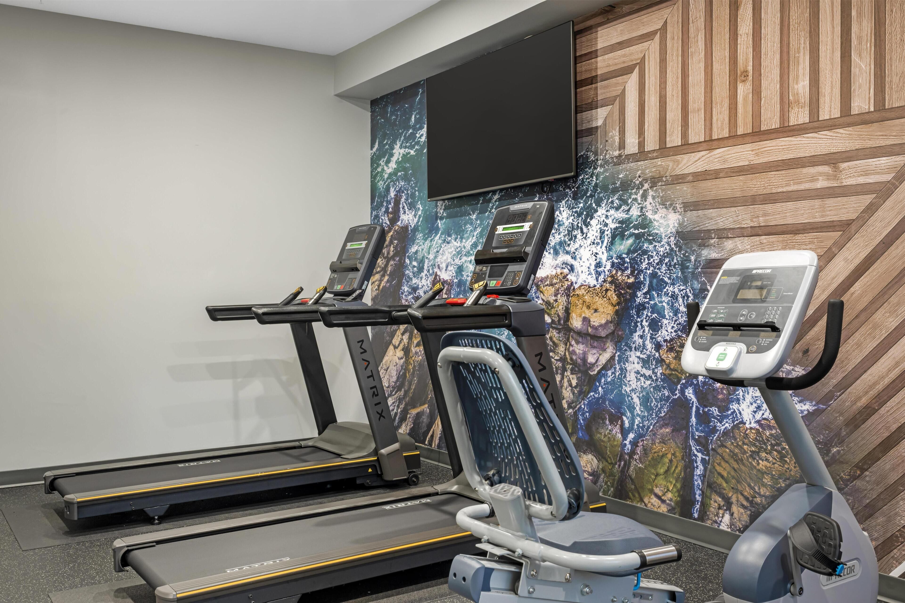 Un gimnasio con cintas de correr, bicicletas estáticas y una pared grande con pantallas.