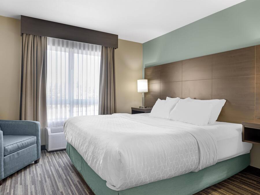 Habitación de hotel con una cama grande, una silla, una mesita de noche, una lámpara y un ventanal con cortinas.