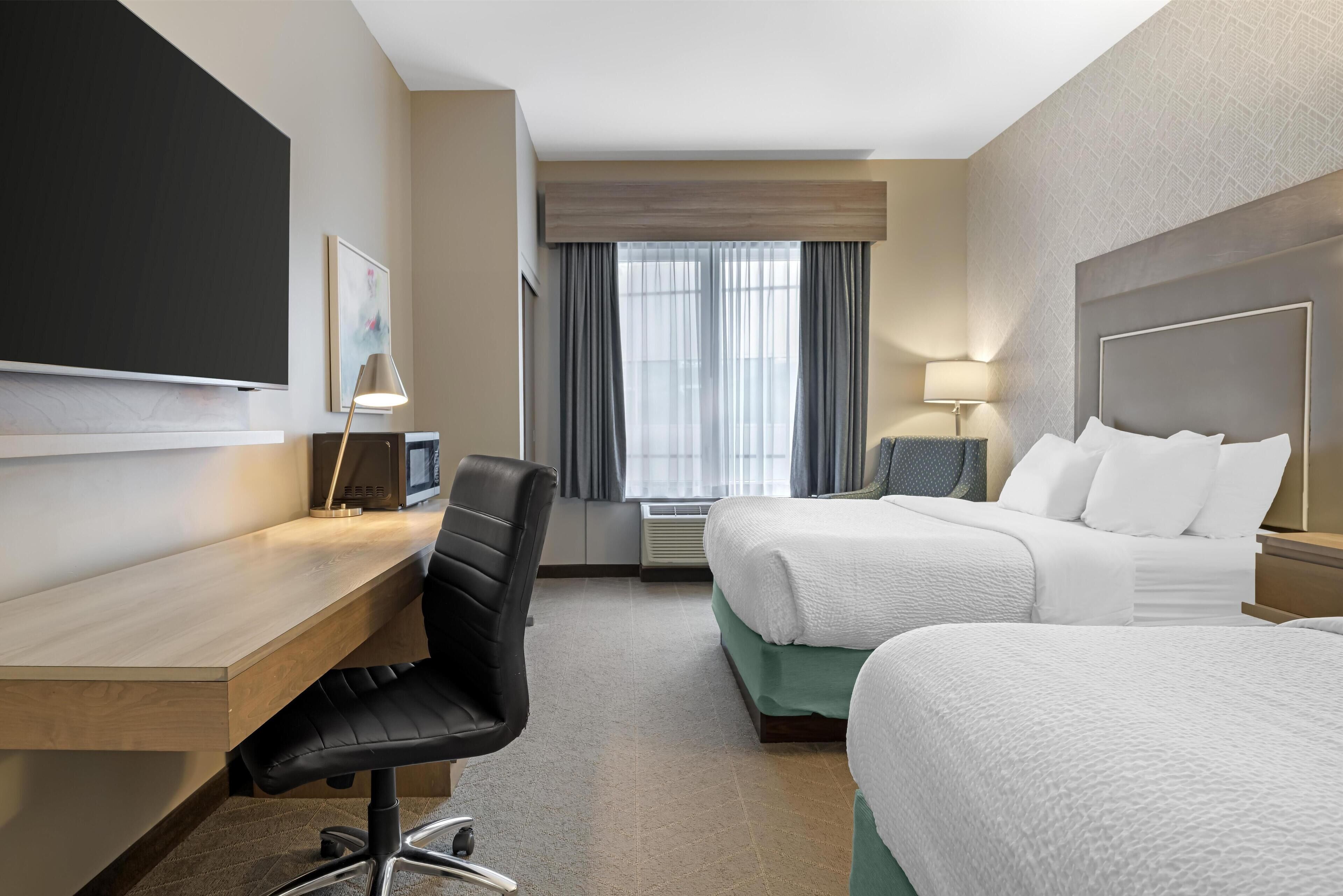 Une chambre d’hôtel avec un bureau en bois, une chaise de bureau noire, un lit avec une literie blanche, une table de chevet avec une lampe et une grande fenêtre avec des rideaux.