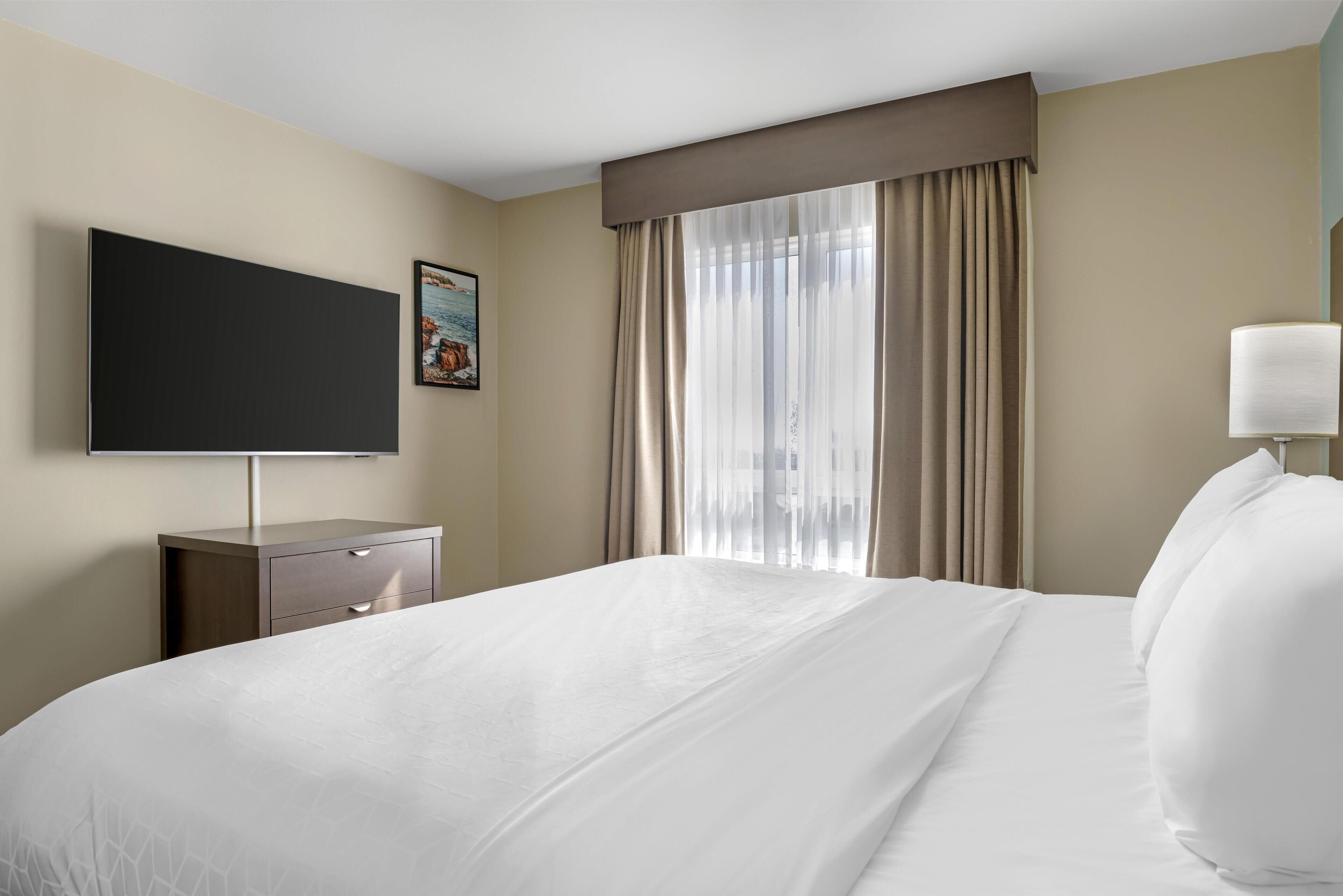 A hotel room with a large bed, a flat-screen TV mounted on the wall, a bedside table with a lamp, and a window with curtains.