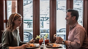 A couple dining at a table with a view of a snowy mountain and ski lift in the background. - Polaris Lodge (Kimberley)