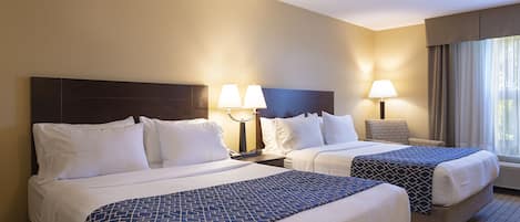 A hotel room with two beds, each with white linens and a patterned blue blanket. There are bedside lamps and a chair near the window.