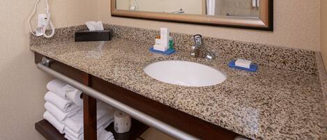 Une salle de bain dotée d’un comptoir en granit, d’un lavabo blanc, d’un porte-serviettes, d’un miroir avec éclairage intégré et d’un sèche-cheveux.