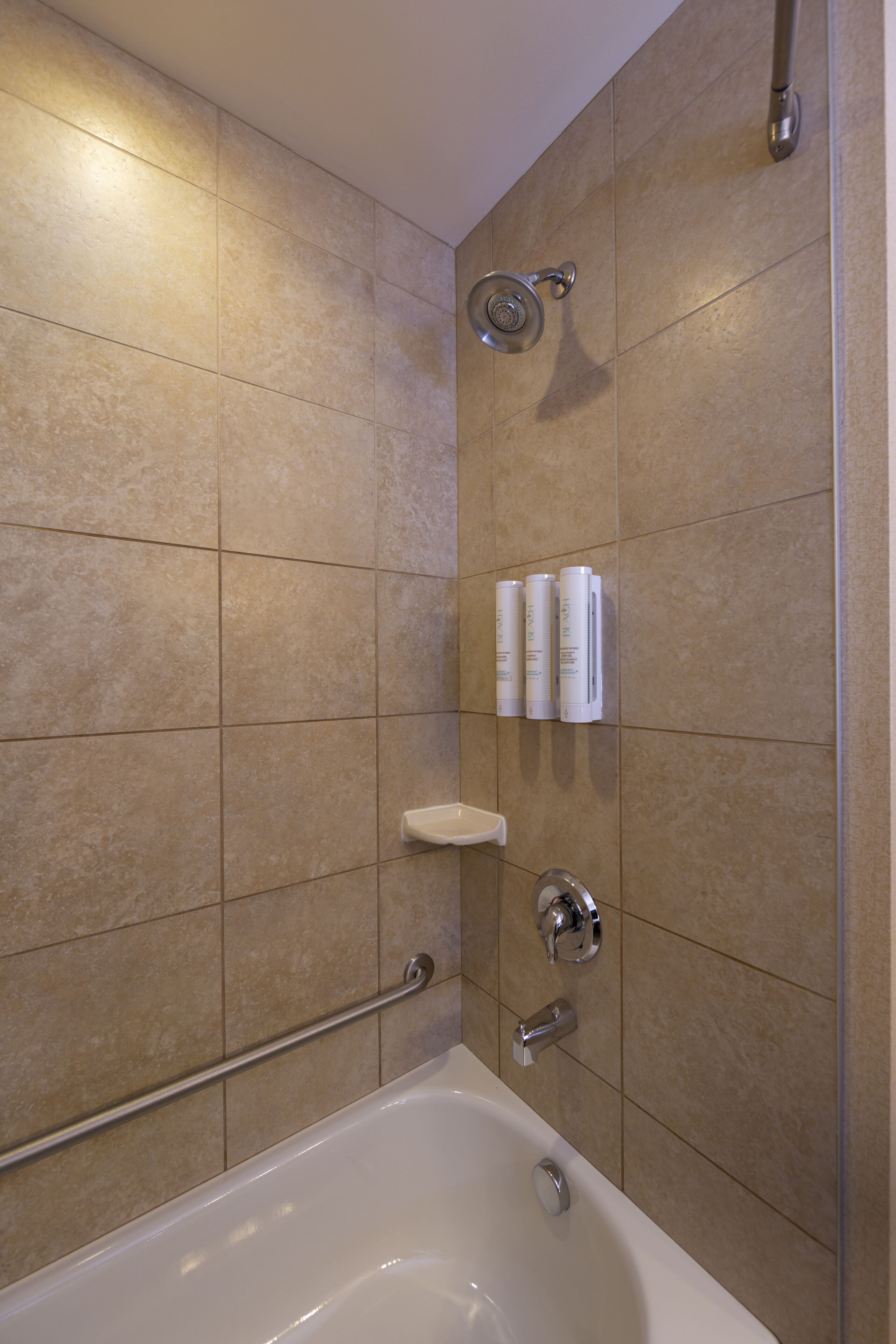 A bathroom with beige tiles, a bathtub, a showerhead, and a handrail.