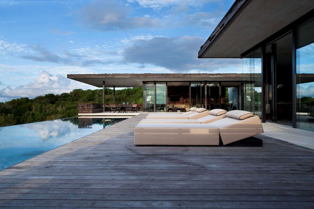 A modern outdoor lounge area with a wooden deck, a pool, and a glass-enclosed space with seating.