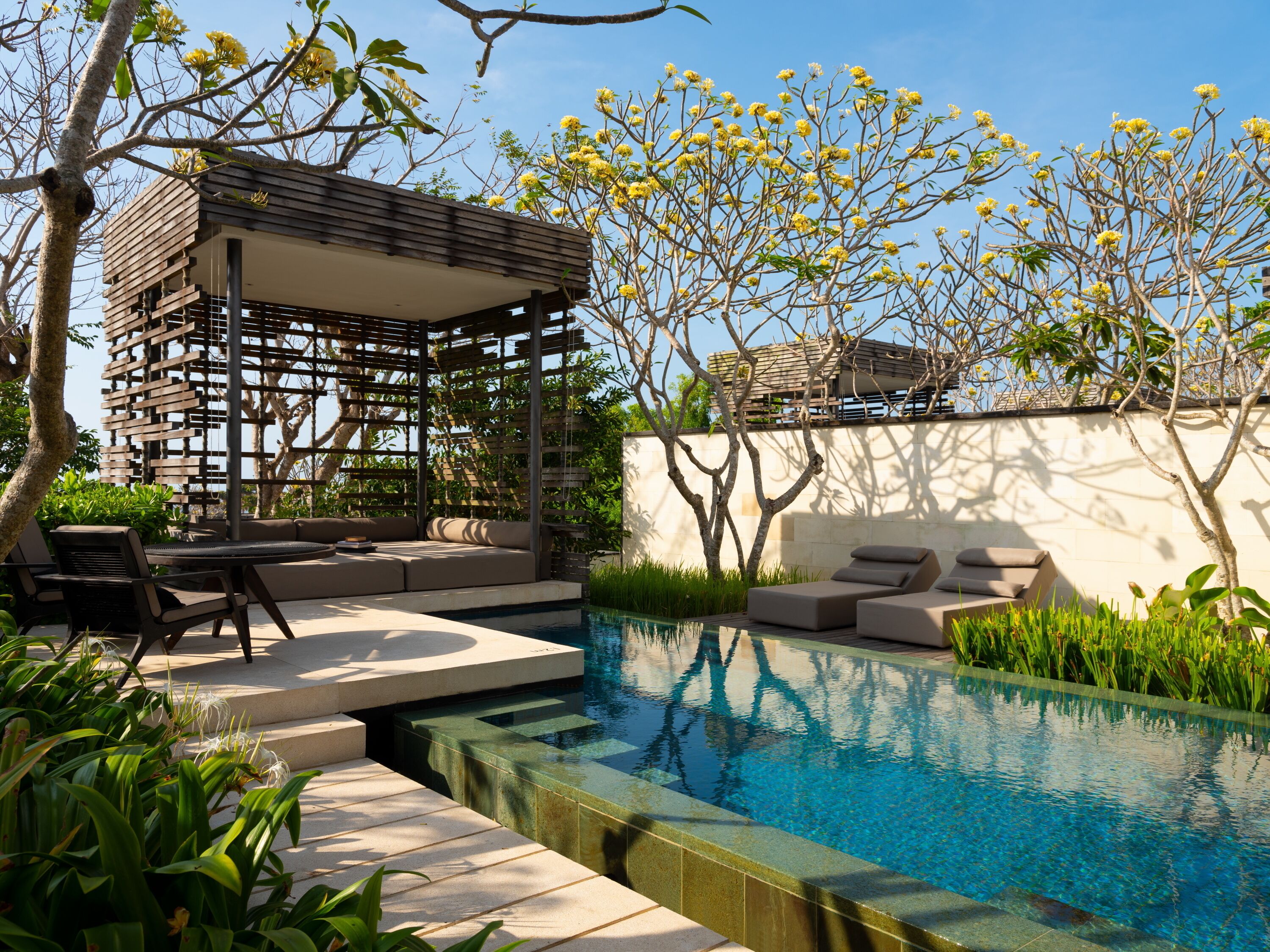 A modern outdoor pool area with a wooden pergola, lounge chairs, and a stone wall.
