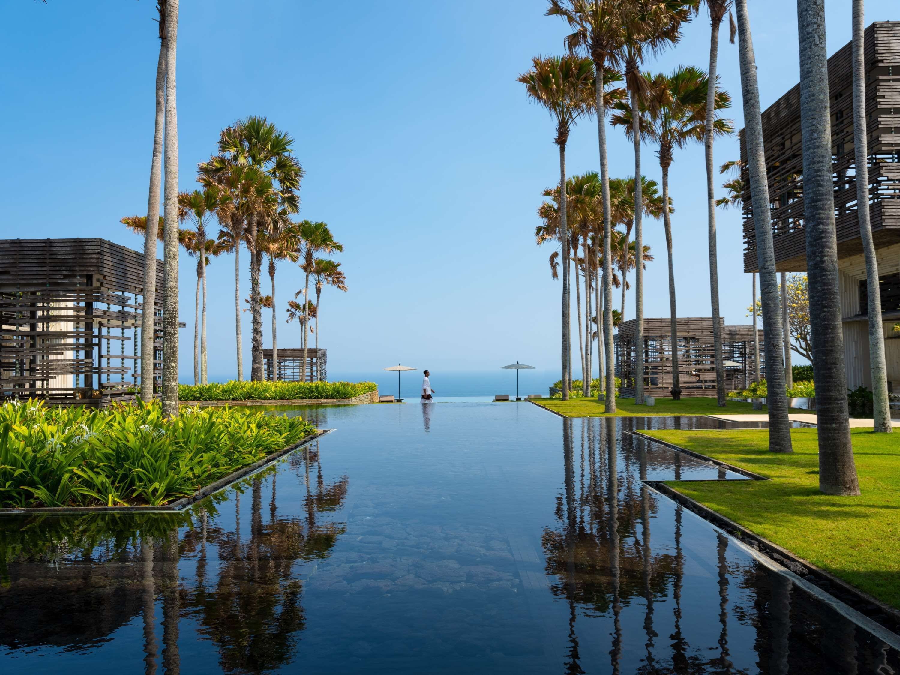 A pool area with palm trees, a person walking, and a view of the ocean.