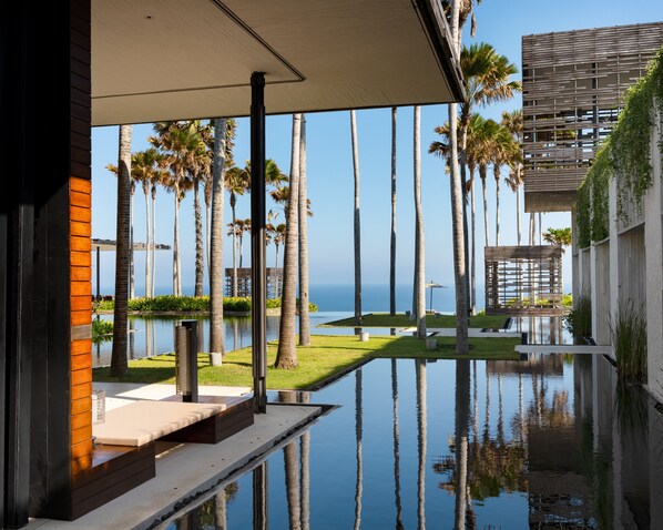 A modern outdoor pool area with a clear view of the ocean, surrounded by tall palm trees and a contemporary building structure.