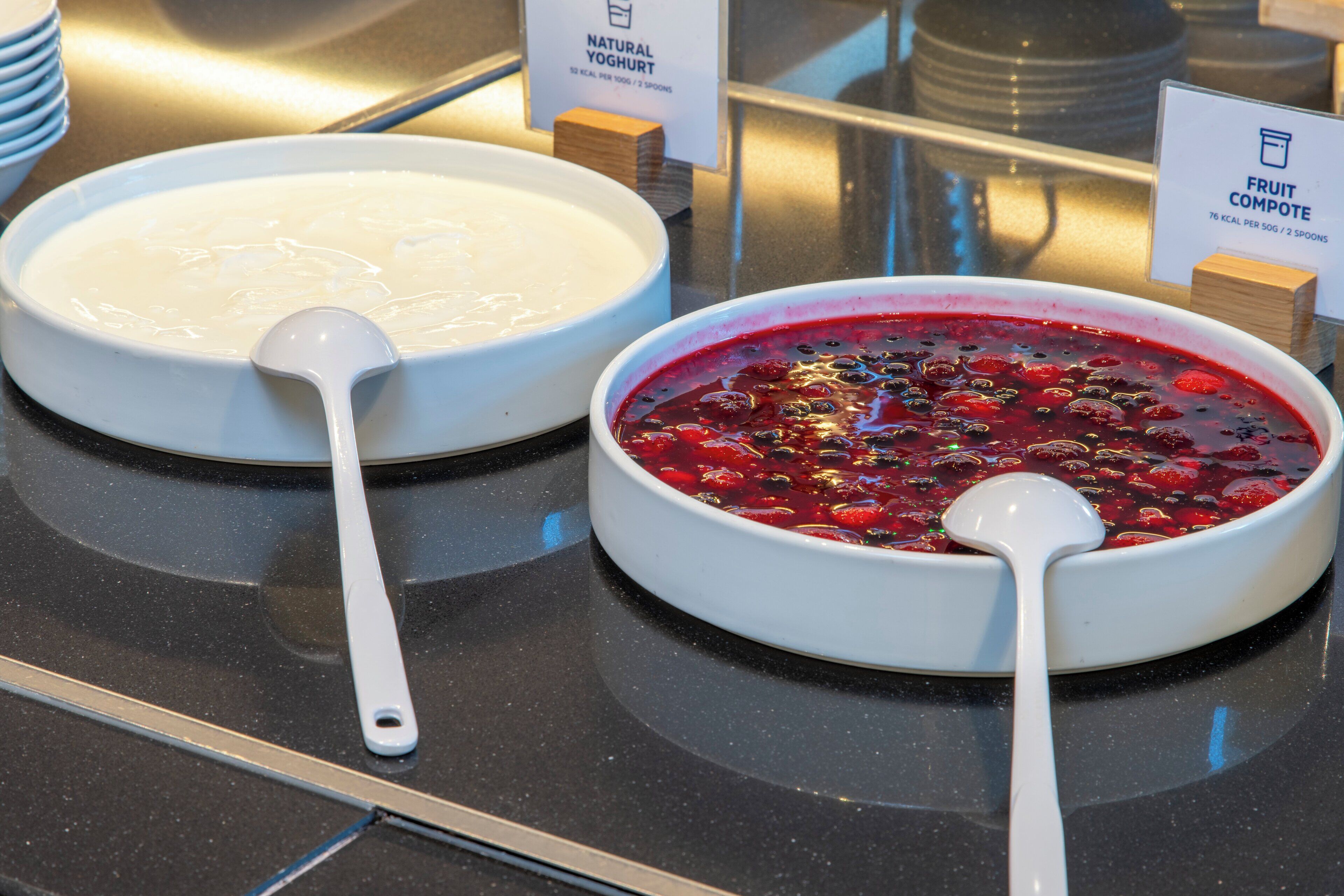 Twee kommen met eten, één met de aanduiding 'Natuurlijke Yoghurt' en de andere 'Vruchtencompote', met lepels erin.