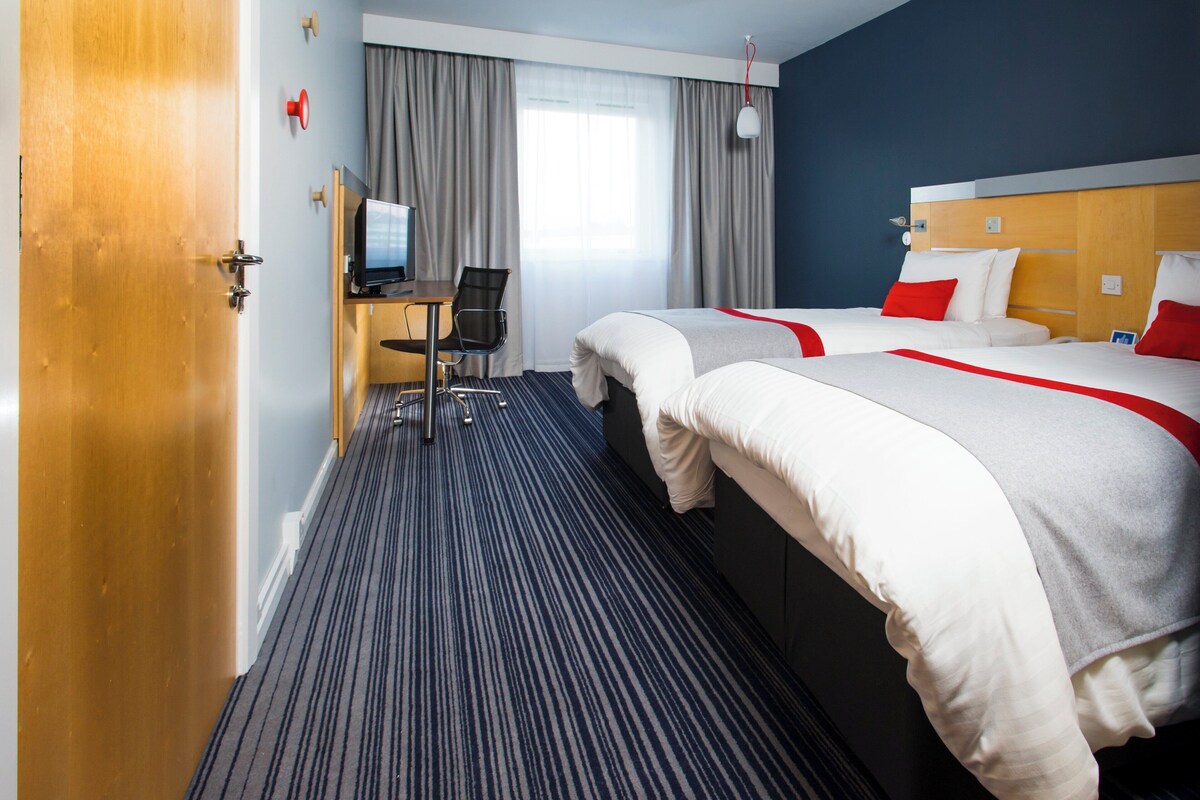 A hotel room with two beds, a desk with a chair, a television, and a window with curtains.