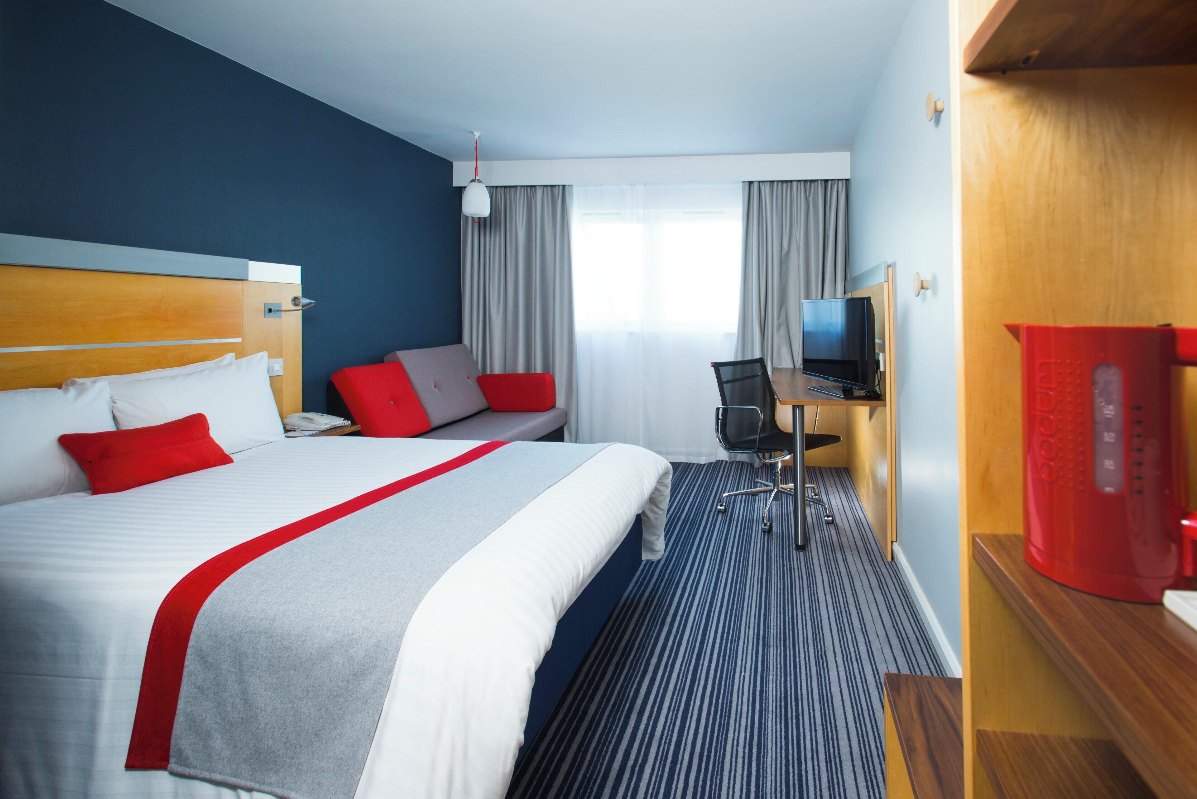 Ein Hotelzimmer mit Bett, Schreibtisch, Sessel, Fernseher und einer kleinen Minibar.